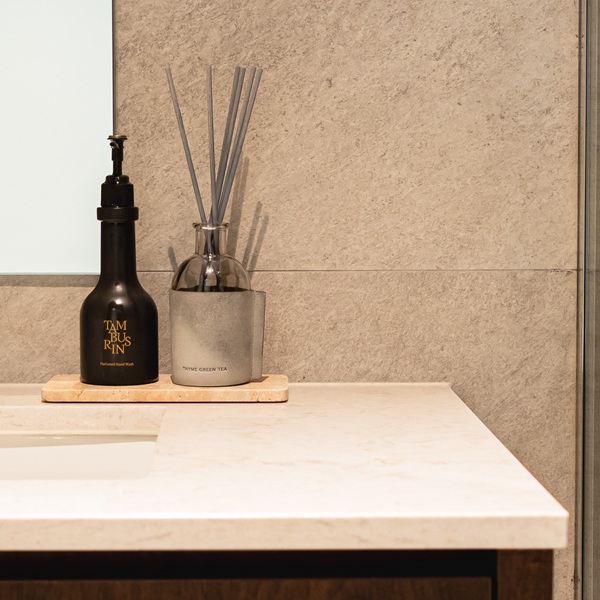 Close-up of an Aurastone vanity top in a common toilet, styled with a black soap dispenser and reed diffuser against a stone-tiled wall.