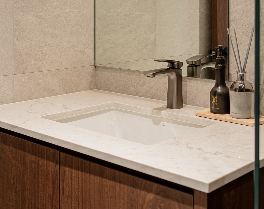 Minimalist bathroom sink with an Aurastone countertop in light beige, paired with wood cabinetry, bronze tapware, and modern accessories.