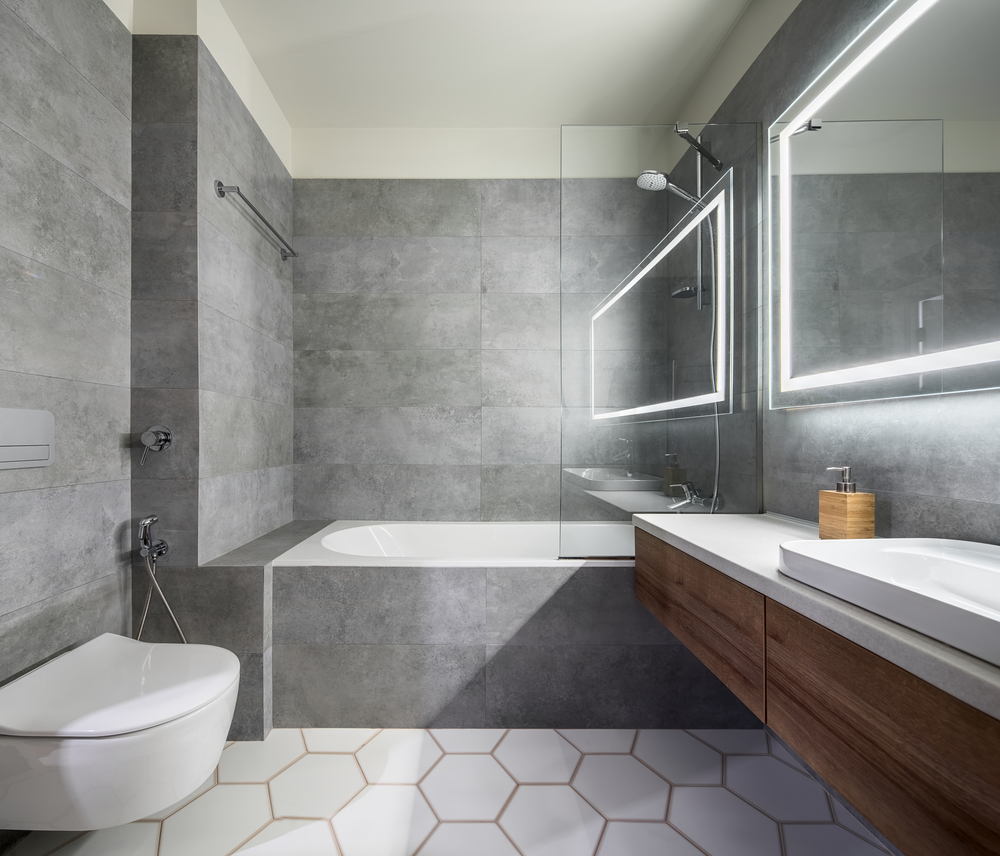 Modern bathroom with gray and white tiles. There is a large mirror with luminous lamps, tabletop with wooden drawers and sink, bath with shower and a glass partition, towel rack and a white toilet. bathroom renovation ideas