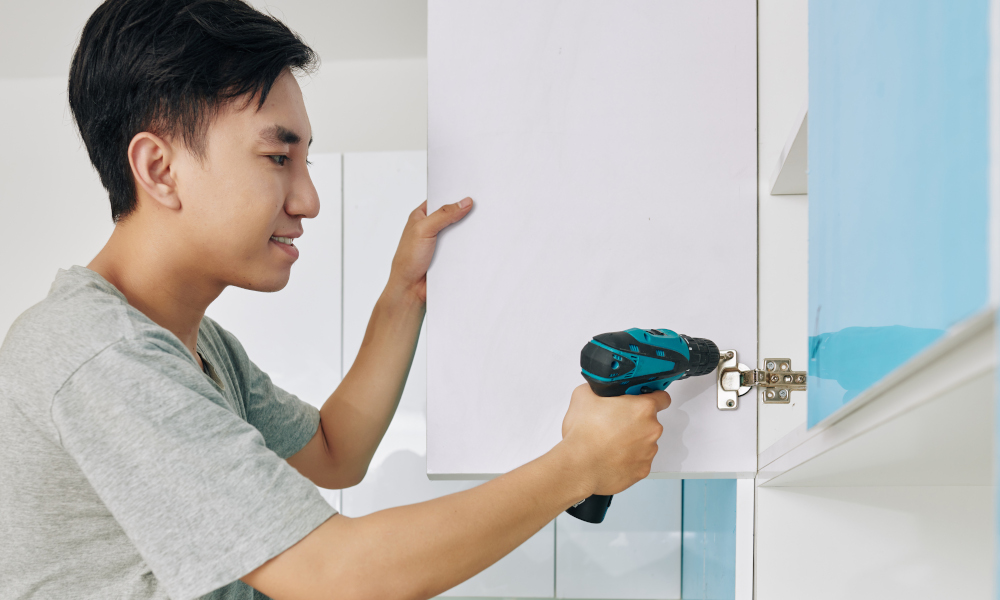 Young man using electric screwdriver when assembling cupboard in kitchen