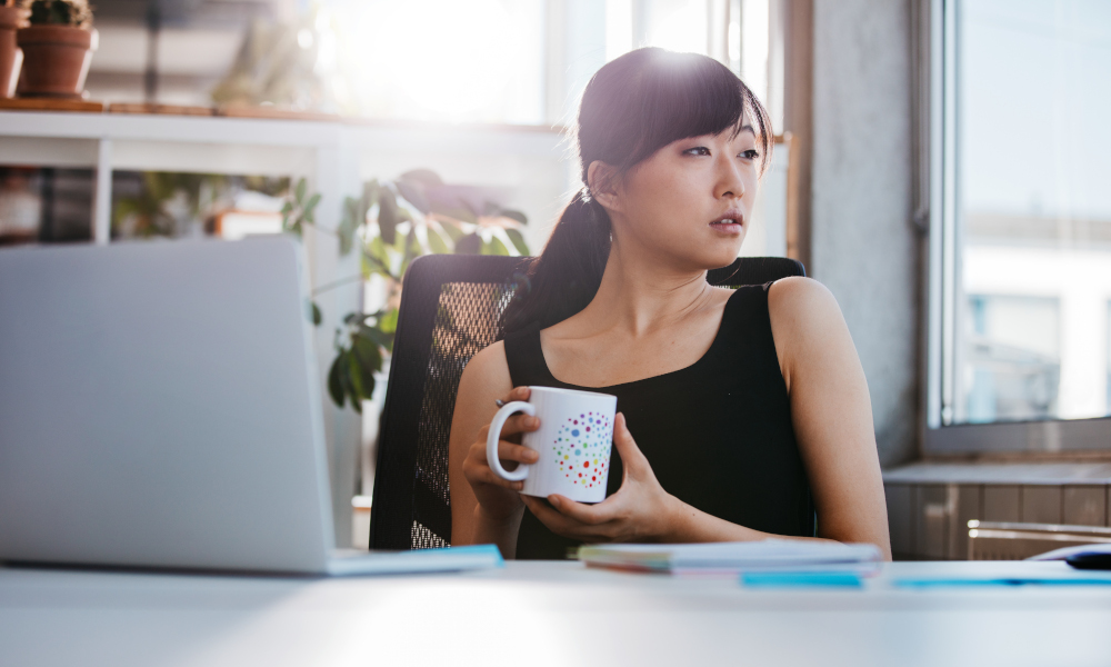 Portrait of relaxed young woman sitting at her desk holding cup of coffee and looking away. Asian business woman taking coffee break in office.