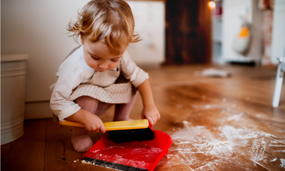 A small toddler girl with brush and dustpan sweeping messy floor in the kitchen at home.