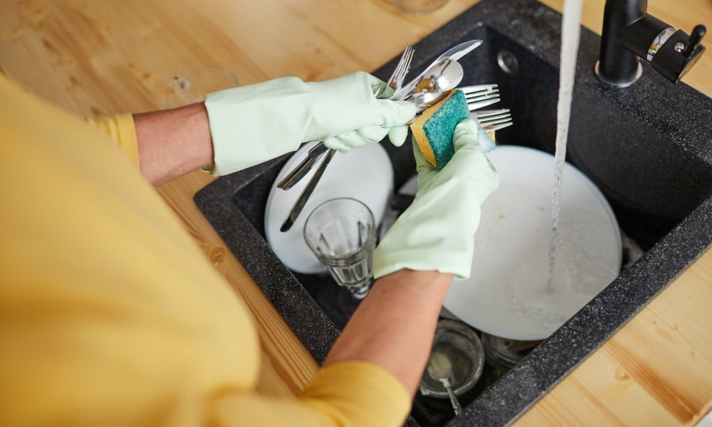 Woman in rubber gloves using sponge while rubbing silverware, she washing dishes in kitchen