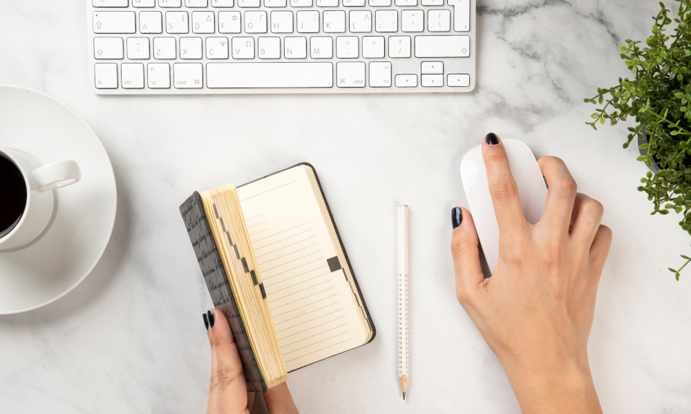 A woman holds a diary and a mouse on a white marble office desk with keyboard and coffee on it