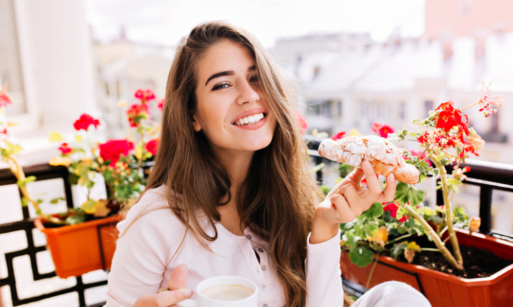 Bbeautiful girl with long hair having breakfast on balcony in the morning in city. She holds a cup, croissant, smiling to camera