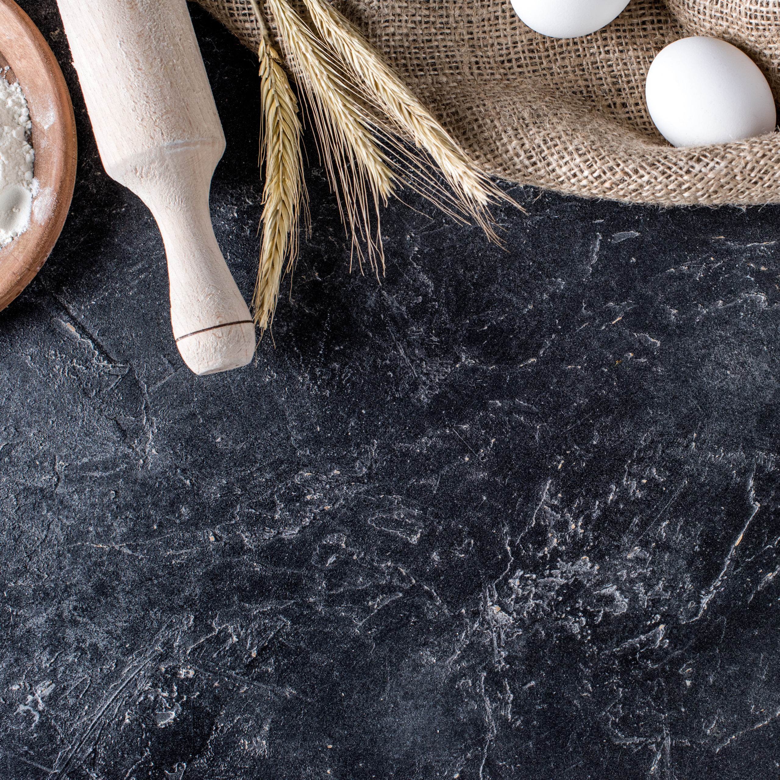 flat lay with wheat, raw eggs on sack cloth and wooden rolling pin on dark marble surface