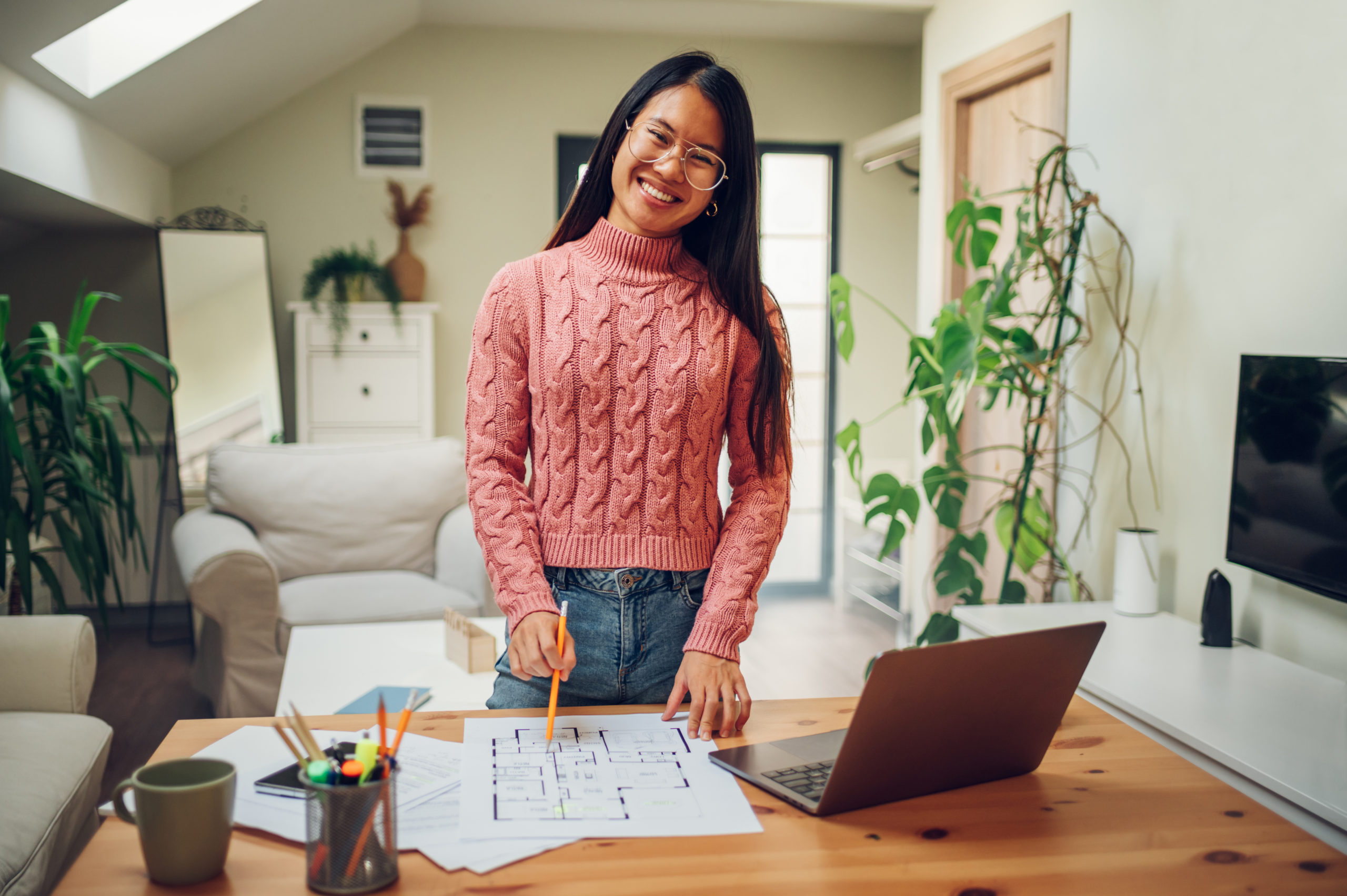 Portrait of smiling Asian female standing at desk at home and studying or working online on a laptop. Happy young Vietnamese woman drawing blueprints or doing paperwork. Looking into the camera.